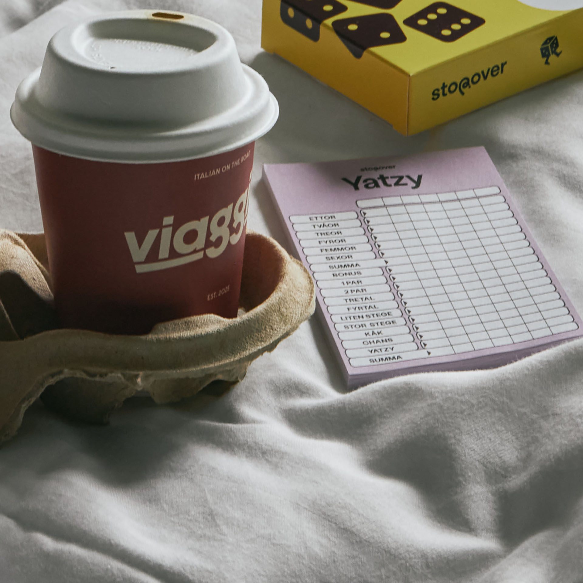 En mugg och Yatzy på en säng. Stopover Hotel