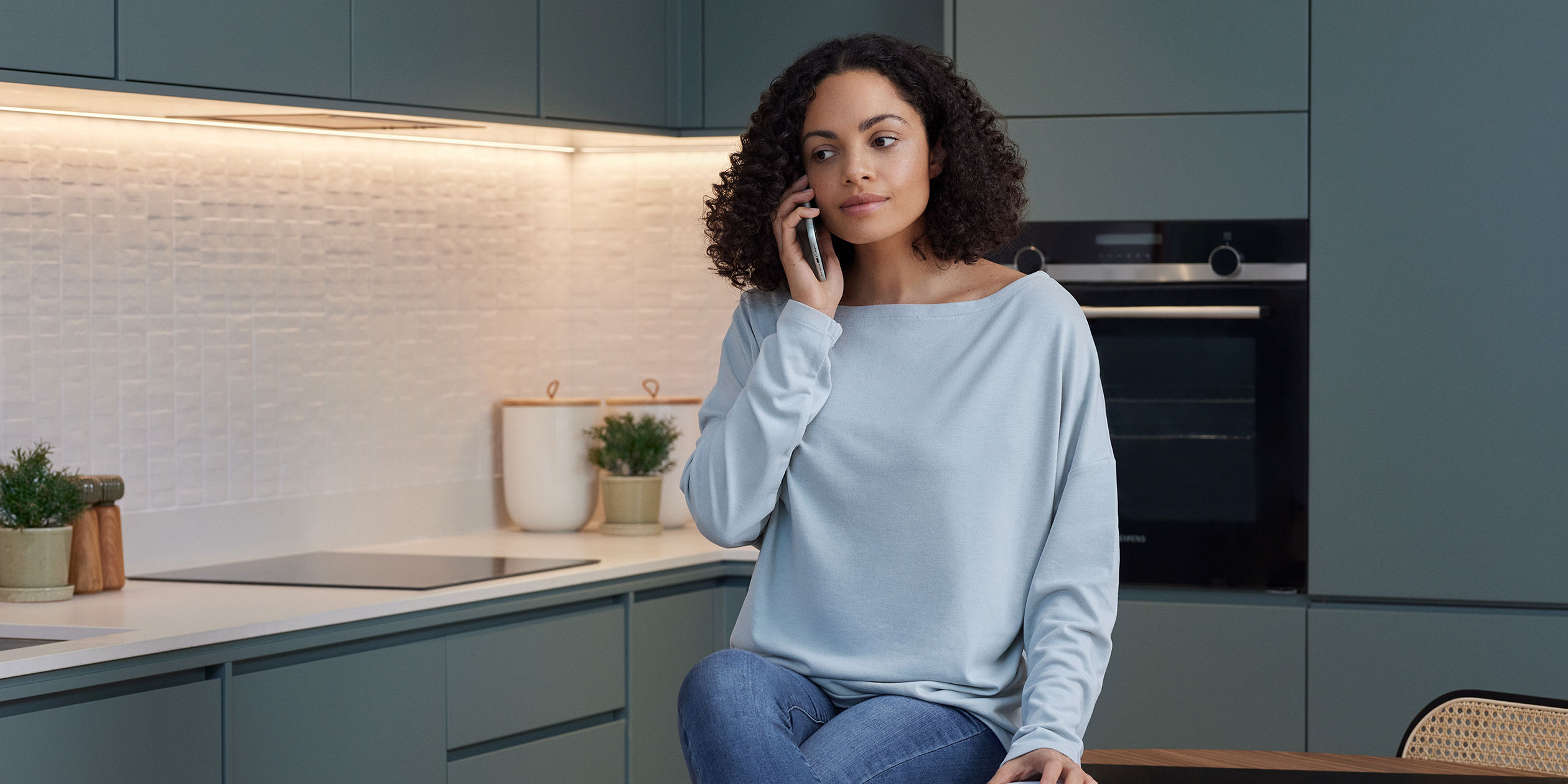 En mörkhårig kvinna pratar i telefon i ett kök.